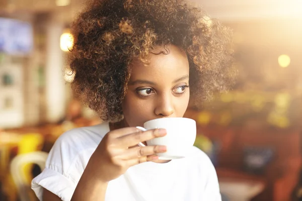Young hipster dark-skinned woman dressed in casual clothes holding cup of hot drink, enjoying coffee or tea. Black student girl spending morning at cafe before going to university, looking thoughtful