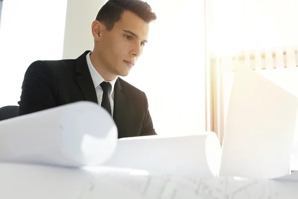 Handsome serious Caucasian contractor in suit working on laptop computer, sitting at desk with blueprints, creating housing project using 3D computer-aided design application, looking concentrated