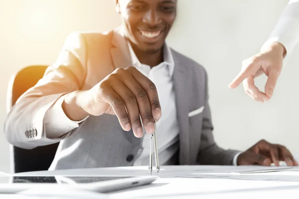 Flare sun. African engineer in stylish gray suit sitting at his desk with copy space tablet and drawings, holding compass, working on new building blueprint while his colleague poiting finger at tool