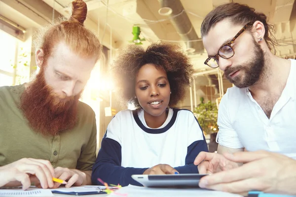 Young entrepreneurs working together at coworking space. Man in glasses holding digital tablet, showing presentation of project to his African female partner while their redhead colleague making notes