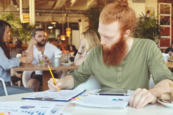 Hipster freelancer holding pencil, making notes in sheets of paper with graphics, using digital tablet for distant work at co-working space. Redhead student doing home assignment at cafeteria