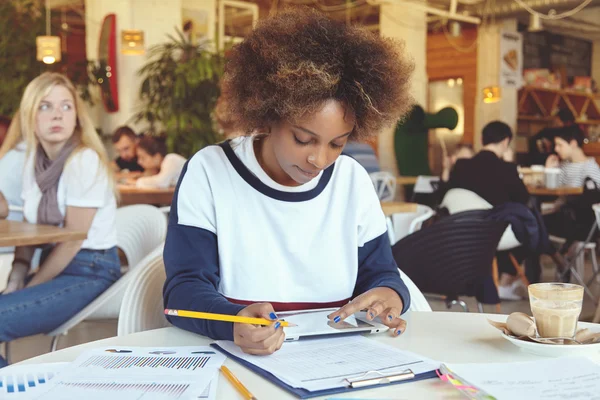 Dark-skinned student girl holding her fingers on touch screen tablet, doing home assignment, preparing for lesson at college canteen, making notes with pencil, looking thoughtful and serious