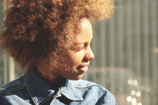 Beautiful African lady with Afro hairstyle and nose-ring, standing outdoors against gray wall with copy space for your text or advertising content, looking aside and squinting her eyes in bright sun