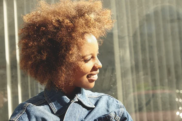 Profile of young fashionable African female with stylish haircut and facial piercing, dressed in denim jacket, looking with joyful expression, standing at wall with copy space for your information