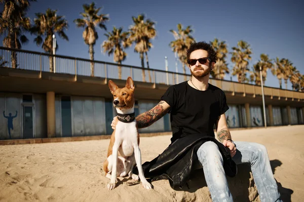 Strong bearded man in sunglasses sitting in sand with friend dog