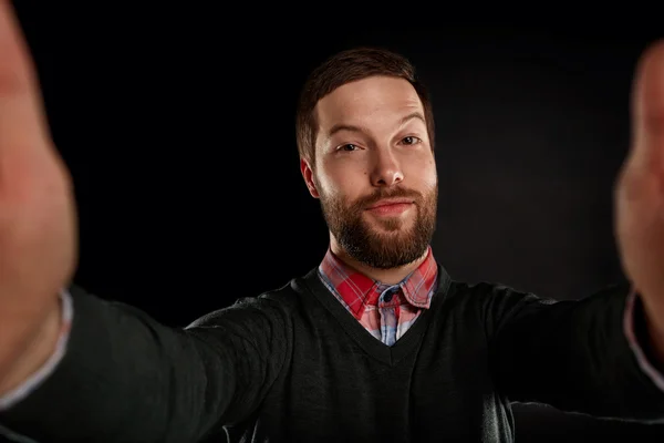 Life style concept. Young man with a beard  in shirt and pullover holding mobile phone and making photo of himself while standing against black background.