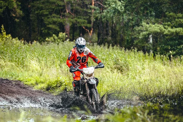 Motorcycle racer rides in a puddle of mud in woods around him water splashes during Ural Cup in Enduro