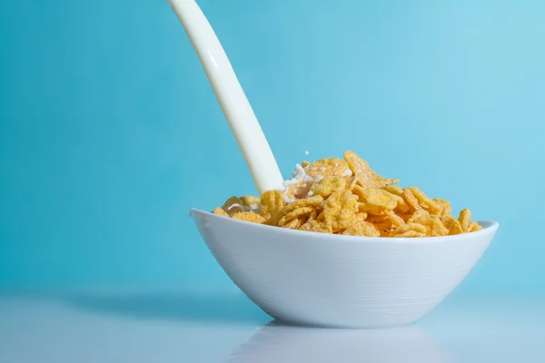 Milk stream pouring into a bowl with yellow flakes, milk splashes on a light blue background