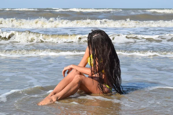 The girl in a yellow bathing suit on the beach. Girl with black hair sitting in sea water head bowed.
