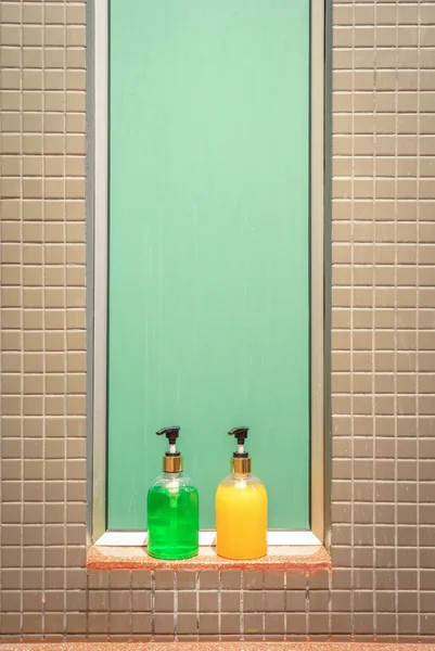 Green and yellow bottles of soap and shampoo against green glass and tiled wall