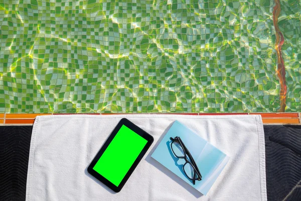 Tablet with blank screen and blue book with glasses lying on white towel on the poolside.