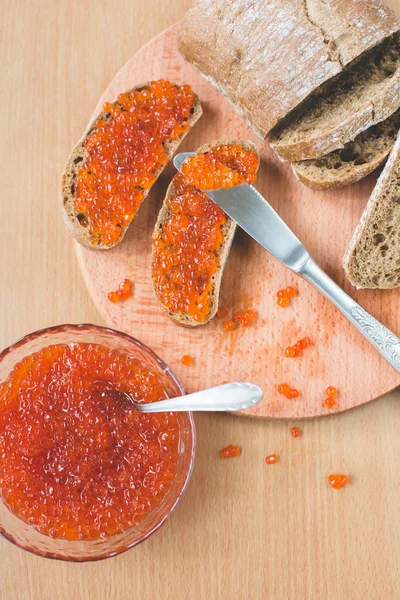 Red caviar toasts sandwich with silver knife, bowl of tasty red caviar with spoon and fresh baked homemade healthy bread on a wooden table background