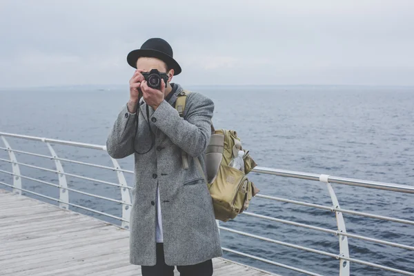 Handsome young hipster guy in hat taking picture with film camera of the sea