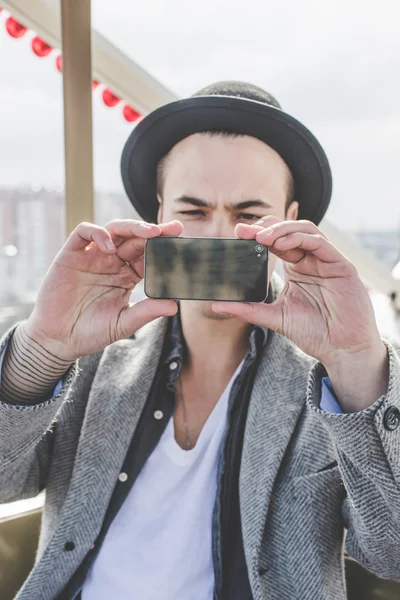 Handsome young hipster guy in hat taking selfie with his smart phone in cabin of ferris wheel over city view from high background during sunny summer day on the festival