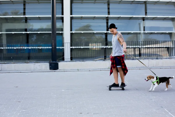 Cool young and handsome caucasian brunette hipster skater guy wearing a hat posing smiling and having fun outside while skating with his skateboard during amazing summer day in the city with his beagle pupppy dog