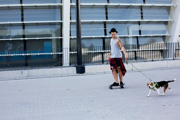 Cool young and handsome caucasian brunette hipster skater guy wearing a hat posing smiling and having fun outside while skating with his skateboard during amazing summer day in the city with his beagle pupppy dog
