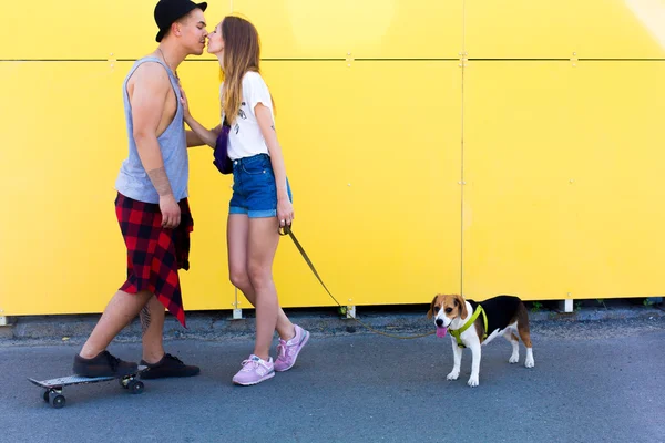 Cool young and beautiful caucasian blonde hipster girl wearing denim shorts with her handsome boyfriend on skate wearing a hat and beagle puppy dog posing smiling kissing having fun and skating outside as a loving perfect hipster couple