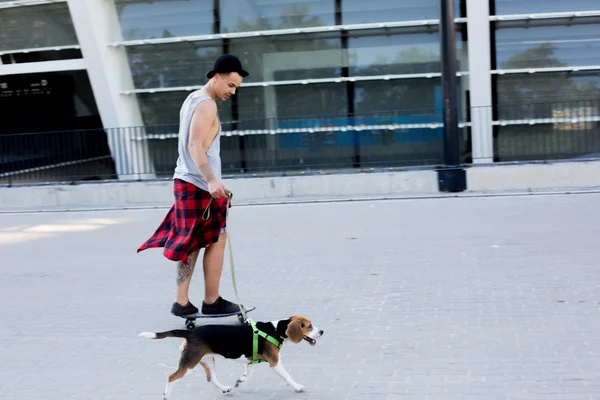 Cool young and handsome caucasian brunette hipster skater guy wearing a hat posing smiling and having fun outside while skating with his skateboard during amazing summer day in the city with his beagle puppy dog