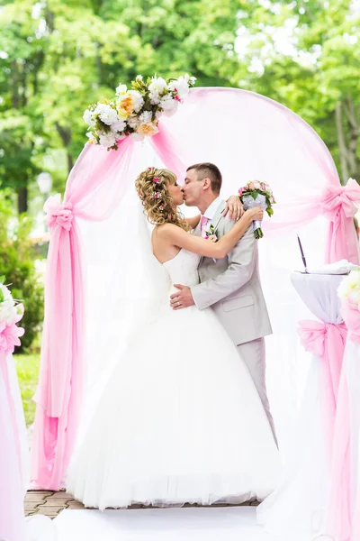 Happy newlywed romantic couple kissing at wedding aisle with pink decorations and flowers
