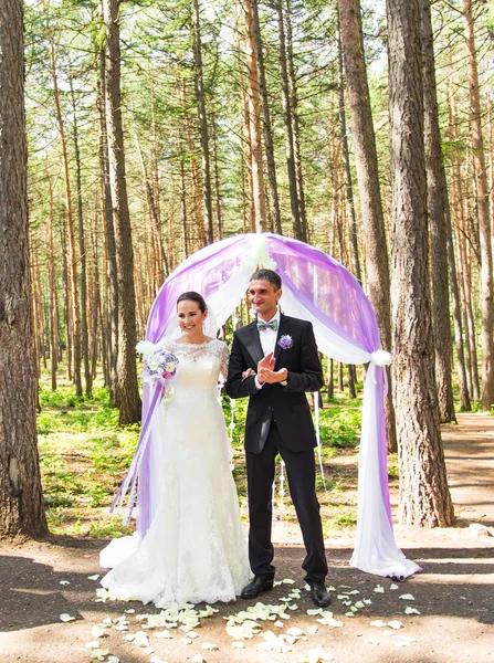 Wonderful stylish rich happy bride and groom standing at a wedding ceremony in green garden near purple arch with flowers