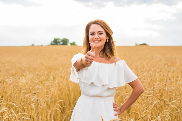 Beautiful young woman giving you thumbs up and smiling in a field with blue sky.