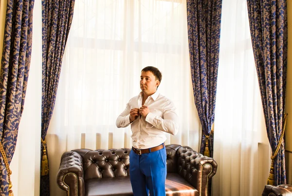 Handsome man putting on shirt standing near window at his room in morning. New opportunities, dating, wedding day or getting ready for job interview concept