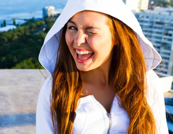 Close up sunset fashion portrait of young teenage girl and sitting on the roof with amazing sea view. Sensual cute fresh happy face, long colored hair, crazy positive emotions. Wearing white hoodie.