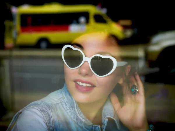 Close up fashion portrait of a beautiful young woman ls sitting at the glass window of street cafe. Stylish romantic trendy girl looking through a big window on traffic jam street. Jeans outfit. Smile