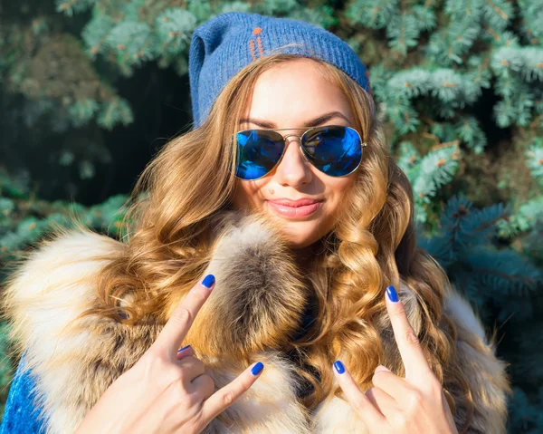 Winter fashion close up winter portrait of Young blonde woman wearing fur vest,blue sunglasses and blue headwear.Excited sexy blonde girl.Looking at camera.doing rock symbol.Outdoors.Luxury concept.