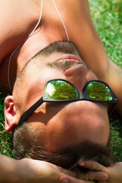 Hipster man relaxed and lying on the grass in city park,in sun reflecting sunglasses,mans fashion style. Close up portrait of Stylish young beard man,sportsman with sexy fit body rest. Summer concept