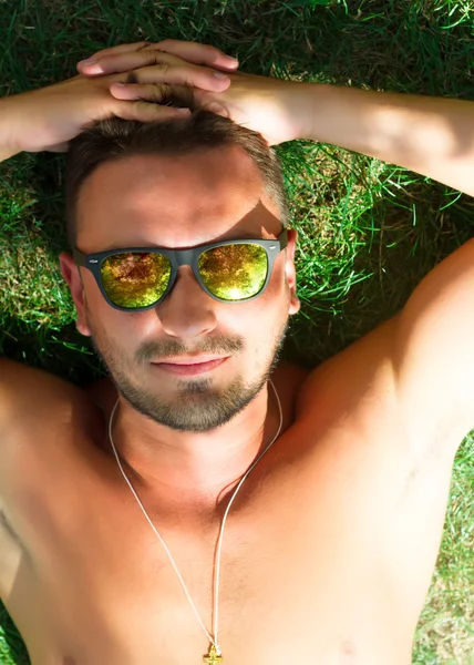 Hipster man relaxed and lying on the grass in city park,in sun reflecting sunglasses,mans fashion style. Close up portrait of Stylish young beard man,sportsman with sexy fit body rest. Summer concept