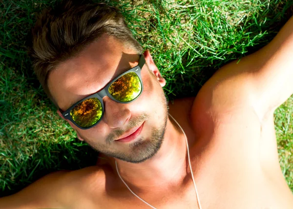 Hipster man relaxed and lying on the grass in city park,in sun reflecting sunglasses,mans fashion style. Close up portrait of Stylish young beard man,sportsman with sexy fit body rest. Summer concept