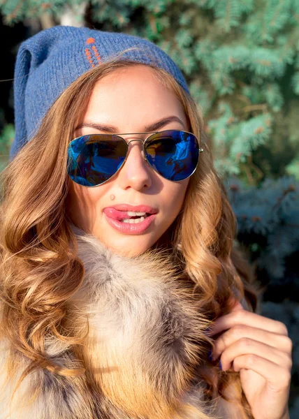 Winter fashion close up winter portrait of Young blonde woman wearing fur vest,blue sunglasses and blue headwear.Excited sexy blonde girl.Looking at camera and showing tongue..Luxury concept.