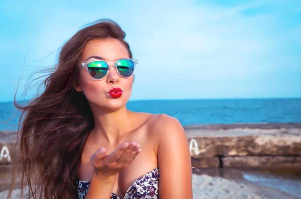 Outdoor summer portrait of young pretty woman looking to the ocean and sending kisses, enjoy her freedom, wearing stylish sunglasses and clothes.