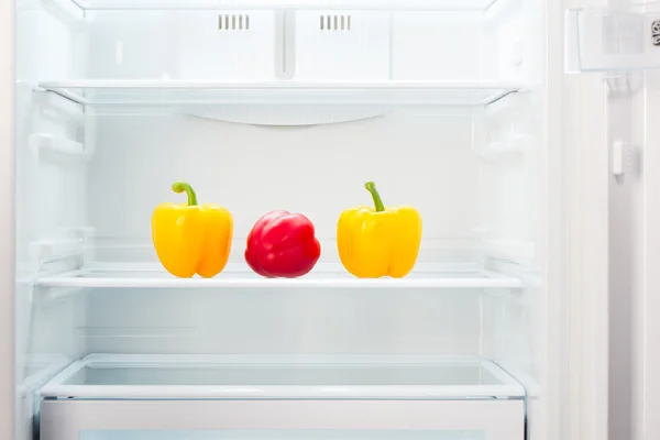 Two yellow and one red peppers on shelf of open empty refrigerator