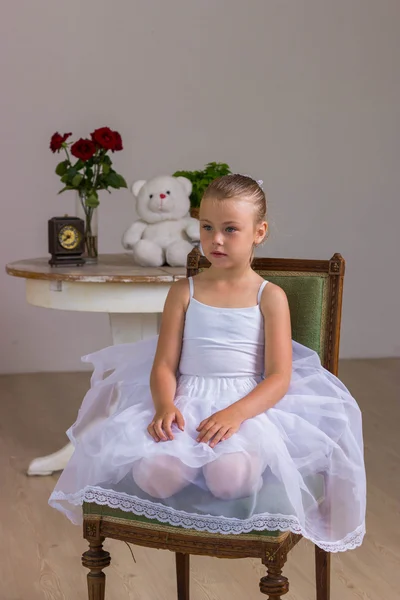 Young ballerina in a white dress sitting on wooden chair on a background with rose and bear