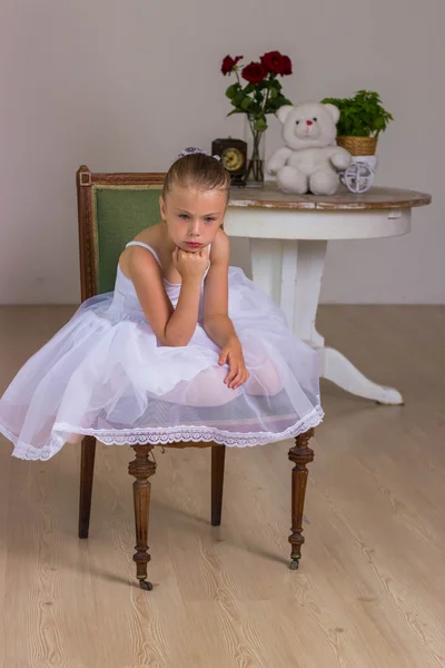 Cute little ballerina in a white dress sitting on wooden chair on a background with rose and bear