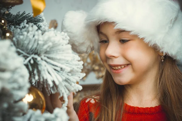 Cute happy girl in red hat hangs decoration on christmas tree