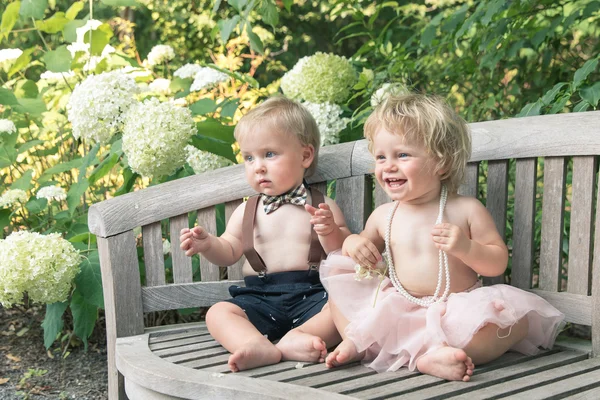 Baby boy and girl in formal dress sitting on wooden bench in a beautiful garden