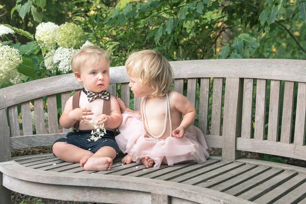 Baby boy and girl in formal dress sitting on wooden bench in a beautiful garden