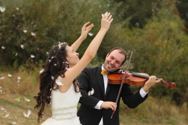 Beautiful bride and groom having fun with feathers in park with groom playing at violin