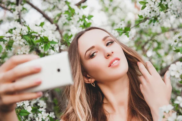 A young girl poses for the camera on the background of a flowering tree. She takes selfie near the flowers.