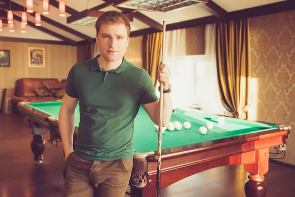 A young man stands in front of a pool table. He looks straight into the camera and holding a cue in his hand. Sunbeams light up the whole room.