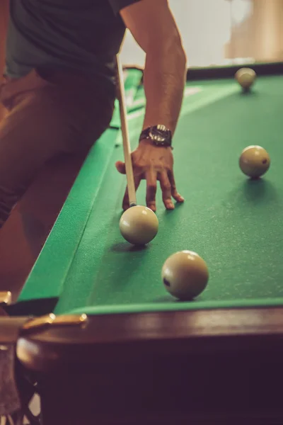 The guy plays billiards and aim the ball in the hole. Focus on center of photo. The guy sitting on the edge of the table.