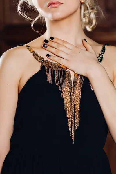 Elegant black dress with gold jewelry on a beautiful sensual young blonde girl with long curly hair and trendy earrings, stylish manicure on nails and luxury make-up holding his hand on his chest