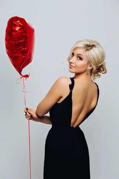 Young beautiful sexy blonde girl with curly hair in an elegant long black dress, standing with a big red heart-shaped helium balloon, isolated on white background trendy makeup
