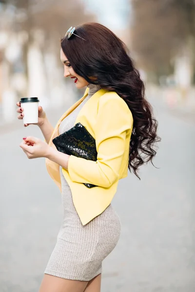 A businesswoman talking via mobile phone and holding a coffee cup. Young slim stylish girl in bright clothes, yellow jacket, a wonderful long dark curly hair holding a cup of coffee and a fashionable