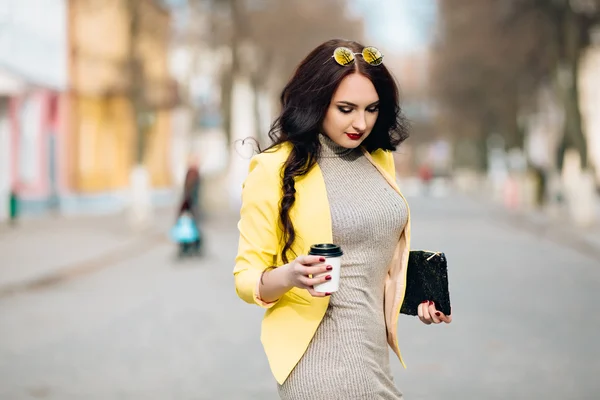 A businesswoman talking via mobile phone and holding a coffee cup. Young slim stylish girl in bright clothes, yellow jacket, a wonderful long dark curly hair holding a cup of coffee and a fashionable