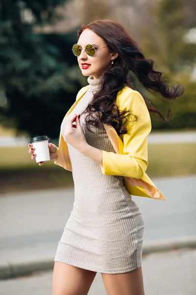 A businesswoman talking via mobile phone and holding a coffee cup. Young slim stylish girl in bright clothes, yellow jacket, a wonderful long dark curly hair holding a cup of coffee and a fashionable