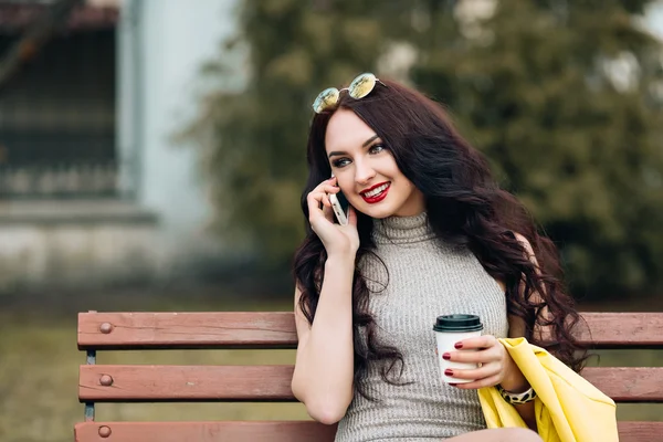 Indoor lifestyle fashion portrait of cute beautiful woman, long curly hair, perfect skin, hipster clothes, yellow shirt, jewelry and sunglasses, drinking coffee, talking on the phone, posing alone.